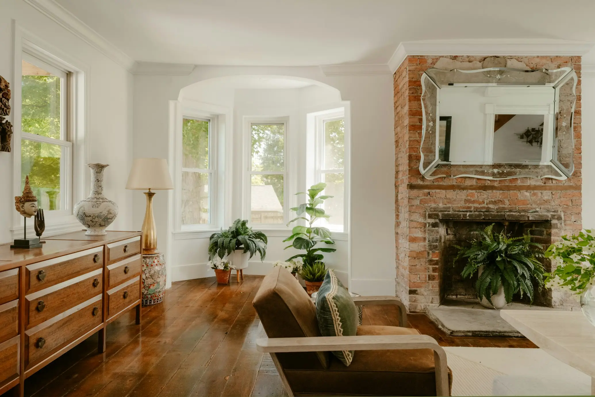 Salon clair avec bow-window, parquet ancien, commode en bois, fauteuil en cuir et cheminée en briques garnie d'une fougère sous un miroir vénitien.